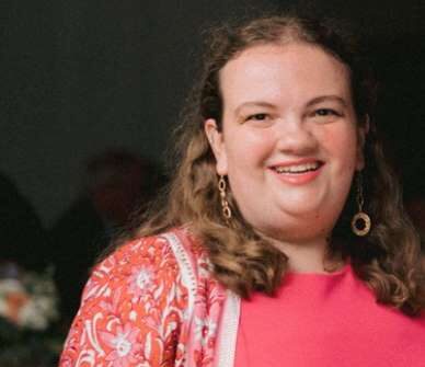 Photo of a woman with shoulder length brown hair wearing a pink shirt and gold earrings.